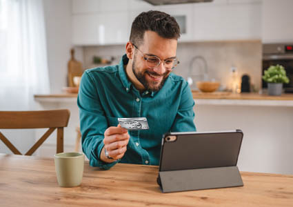 Man sitting at kitchen table holding his EECU Visa credit card and looking on his tablet